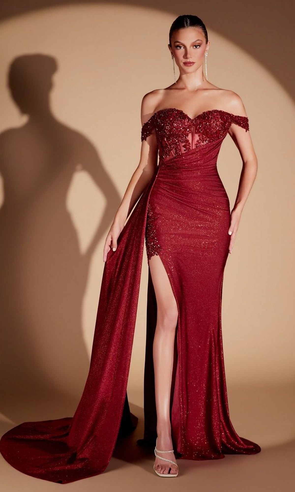 A woman stands against a beige background wearing the Ladivine J914, a burgundy long prom dress featuring an off-the-shoulder, form-fitting silhouette with a thigh-high slit, sweeping train, glittery fabric, elegant sweetheart neckline, and a sequin embroidered corset bodice.